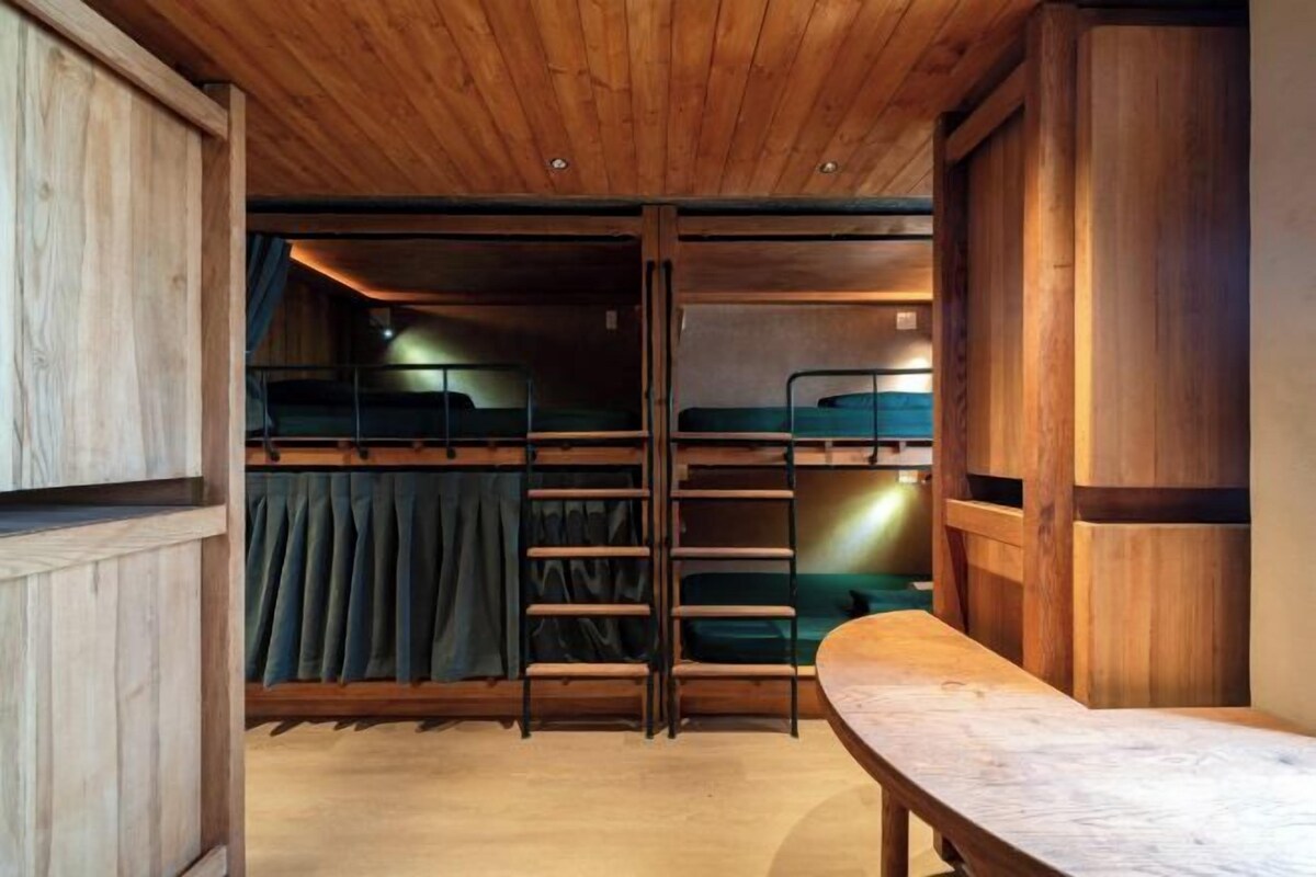 Een slaapkamer met stapelbed, houten plafond, een bankje en een gordijn.