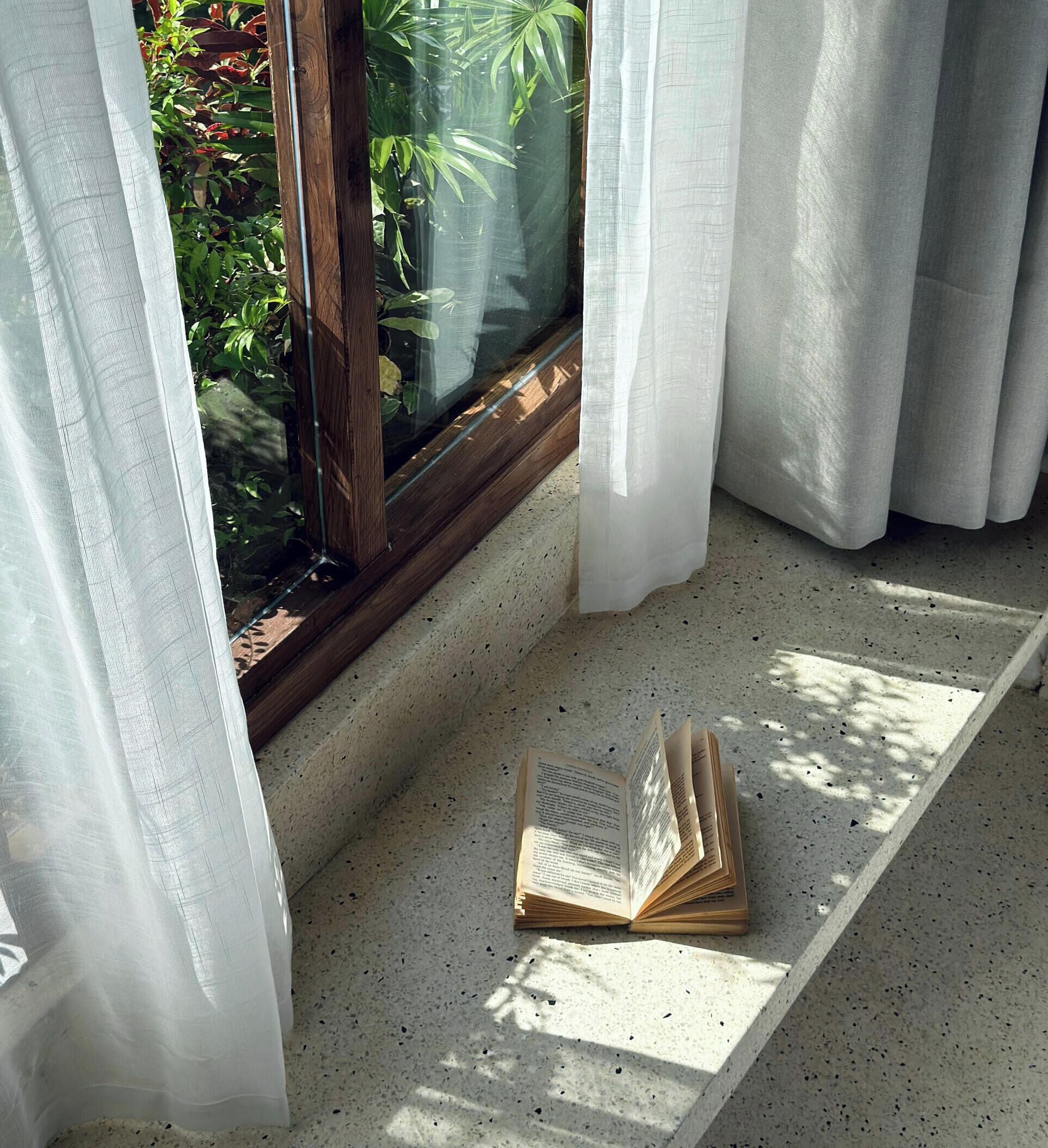 A window with a view of greenery, a book on a ledge, and white curtains.