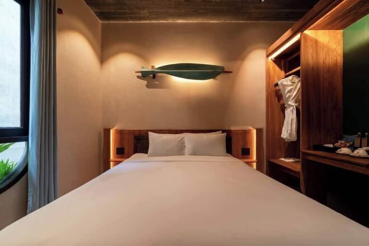 A hotel room with a bed, a wooden headboard, a nightstand, a robe hanging on a wooden rack, and a wall-mounted light fixture.
