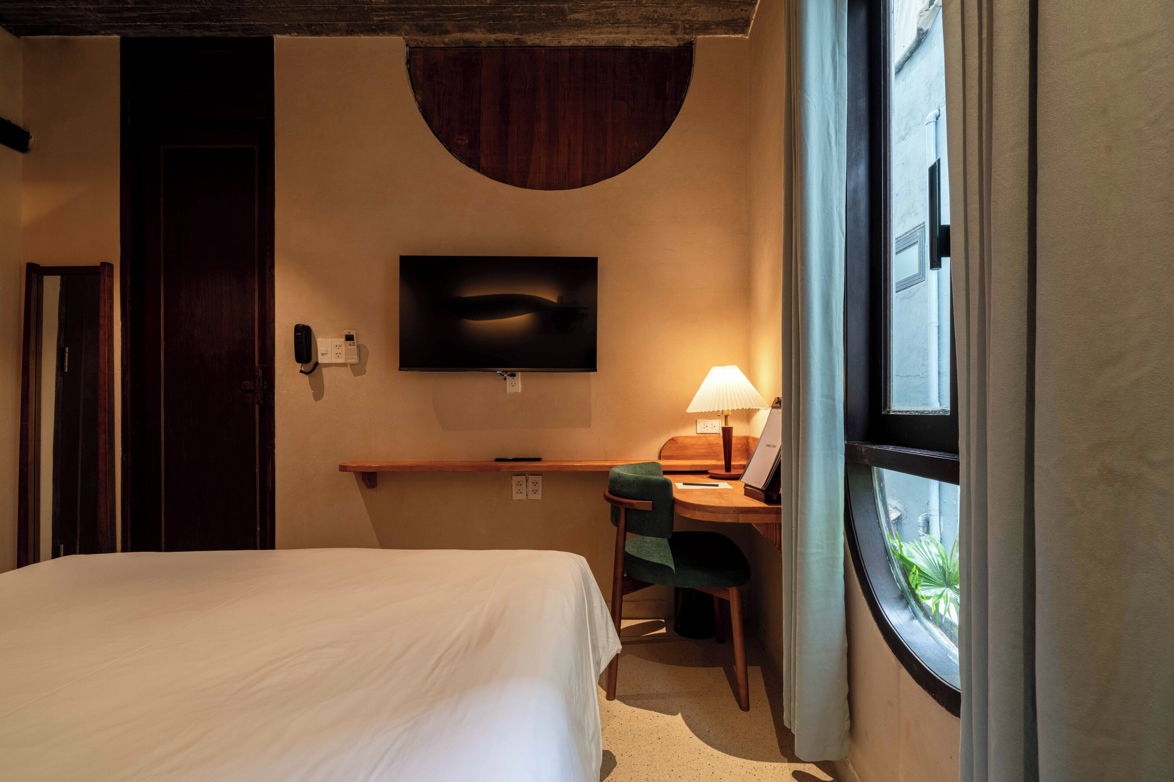 Quarto de hotel com cama, escrivaninha com cadeira, televisão e janela com cortinas.