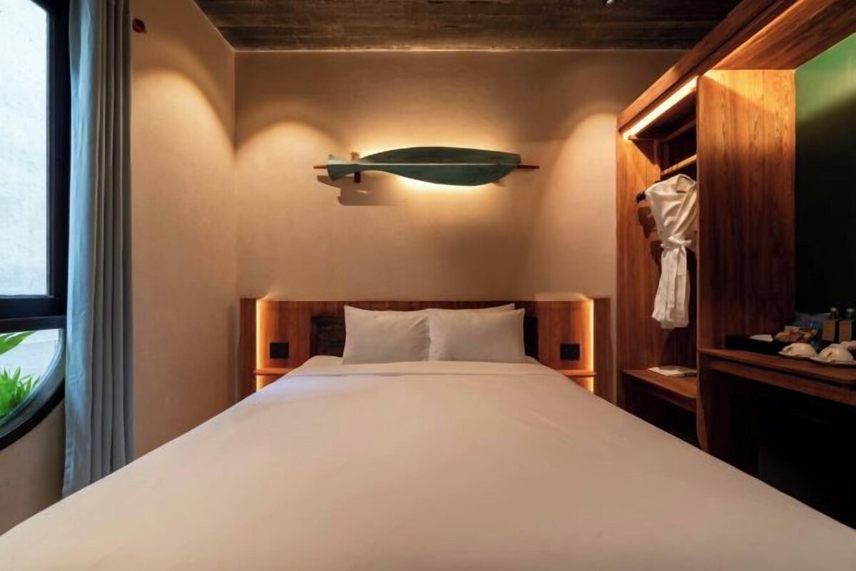 Hotelkamer met een bed, een houten hoofdbord, een nachtkastje, een badjas hangend aan een houten rek en een wandlamp.