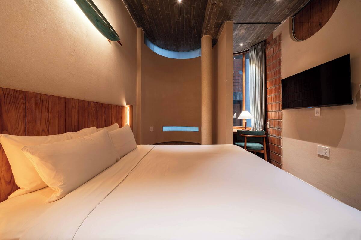 A hotel room with a large bed, a flat-screen TV, a wooden headboard, and a window with curtains.