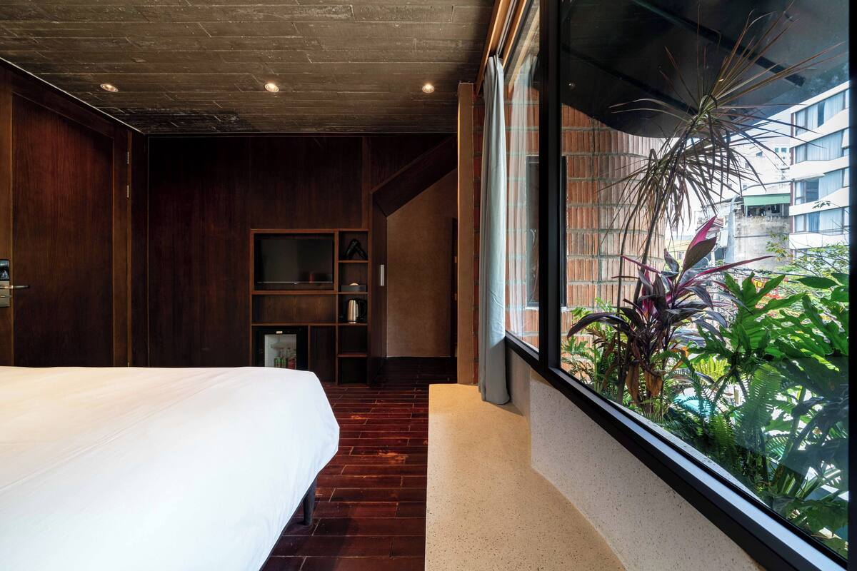 A modern hotel room with a large bed, wooden flooring, a built-in shelf unit, and a view of greenery through a large window.