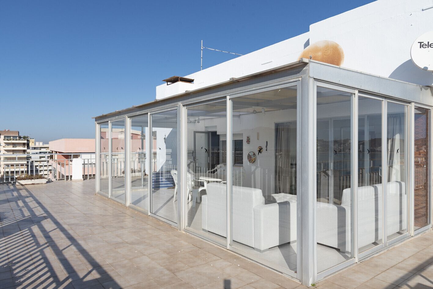 Un área de estar al aire libre con paredes de vidrio, mobiliario blanco, vista despejada de los edificios circundantes y techo plano.