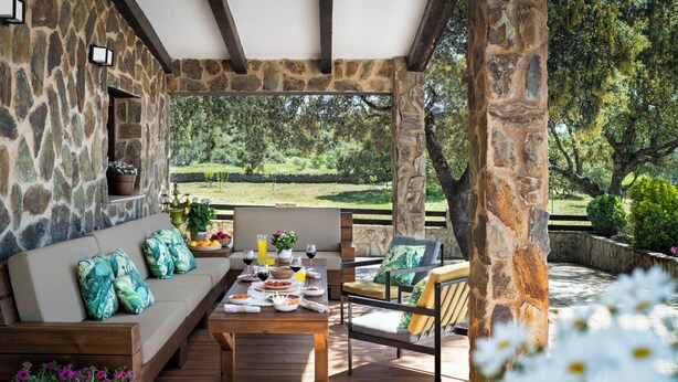 A covered outdoor seating area with stone walls, a wooden table set with food and drinks, and a view of a lush garden. - Rural house surrounded by mountains with swimming pool, skatepark and children's playground in Llerena (Ribera de los Molinos, Llerena)