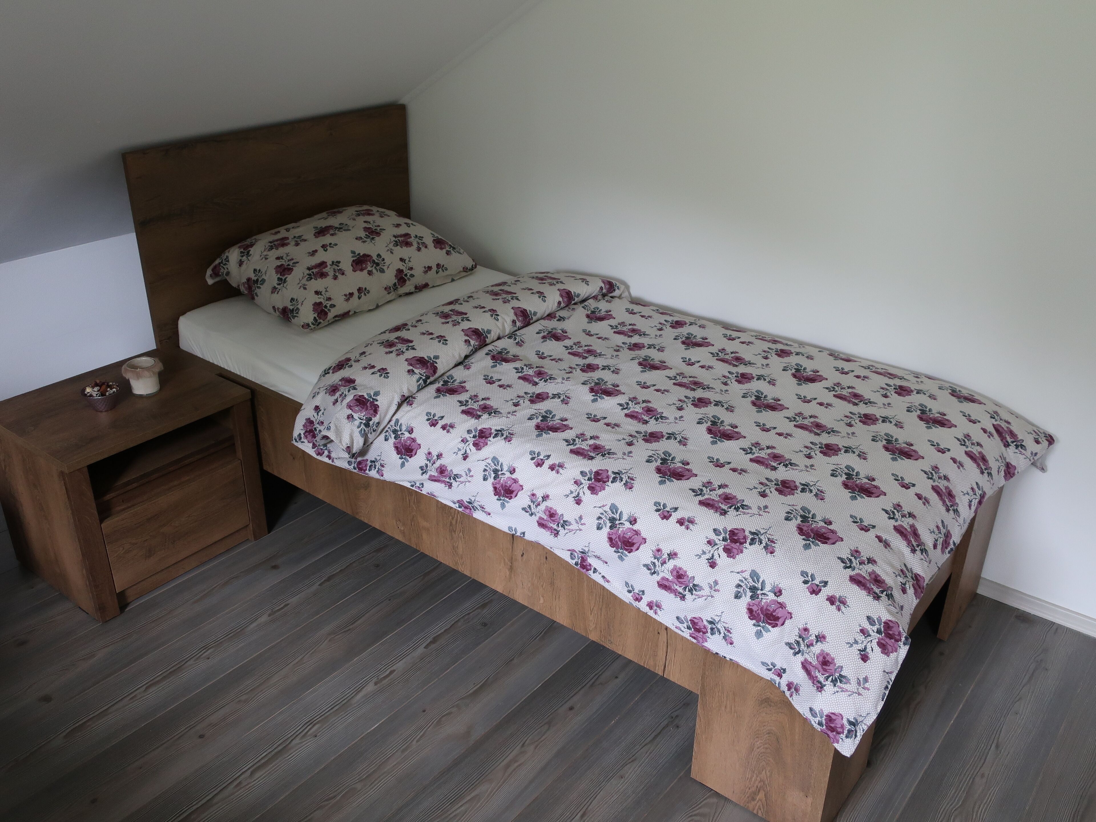 Uma cama de solteiro com colcha florida e almofada combinando, um criado-mudo de madeira com uma tigela e uma vela, e um chão de madeira escura.