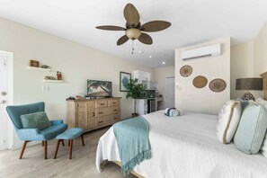Quarto com cama, uma cadeira, cômoda, ventilador de teto e área de cozinha ao fundo.