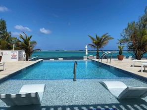 A pool with clear blue water, surrounded by lounge chairs and palm trees, with a view of the ocean. - The One Love Ocean View Studio Cottage in East End (East End)