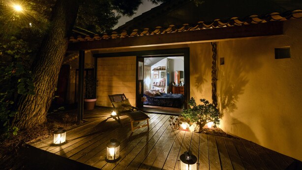 Una terraza de madera con dos sillas y faroles, que da a una habitación con una cama y una mesita.
