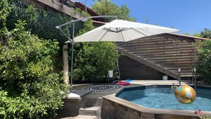 Una terraza de madera con un jacuzzi, rodeada de abundante vegetación y una pérgola con sombrilla.