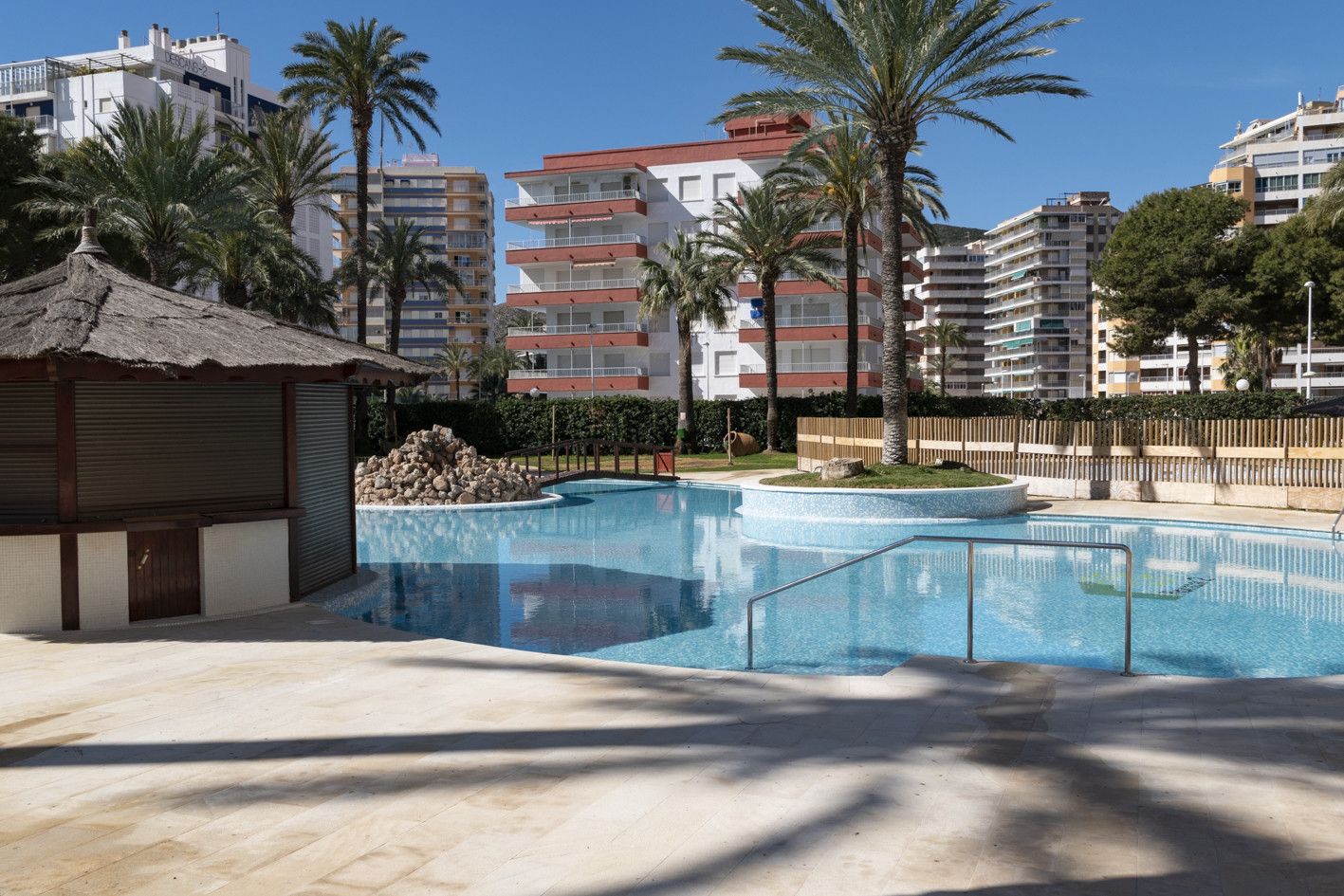 Une piscine dotée d’une structure en pierre, bordée de palmiers, avec des bâtiments modernes en arrière-plan.