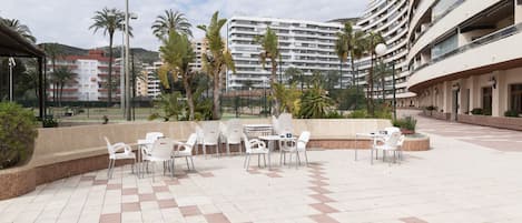 An outdoor patio area with white plastic chairs and tables, surrounded by a low brick wall and palm trees.