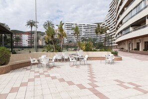 An outdoor patio area with white plastic chairs and tables, surrounded by a low brick wall and palm trees.