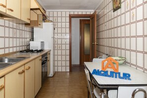 A compact kitchen with a stove, sink, and refrigerator. A dining table with a bowl is visible in the foreground.