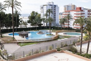 A resort with a central swimming pool, palm trees, and a view of the sea.
