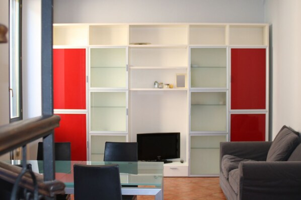 A modern living room with a glass coffee table, a flat-screen TV, a grey sofa, and a red and white shelving unit.