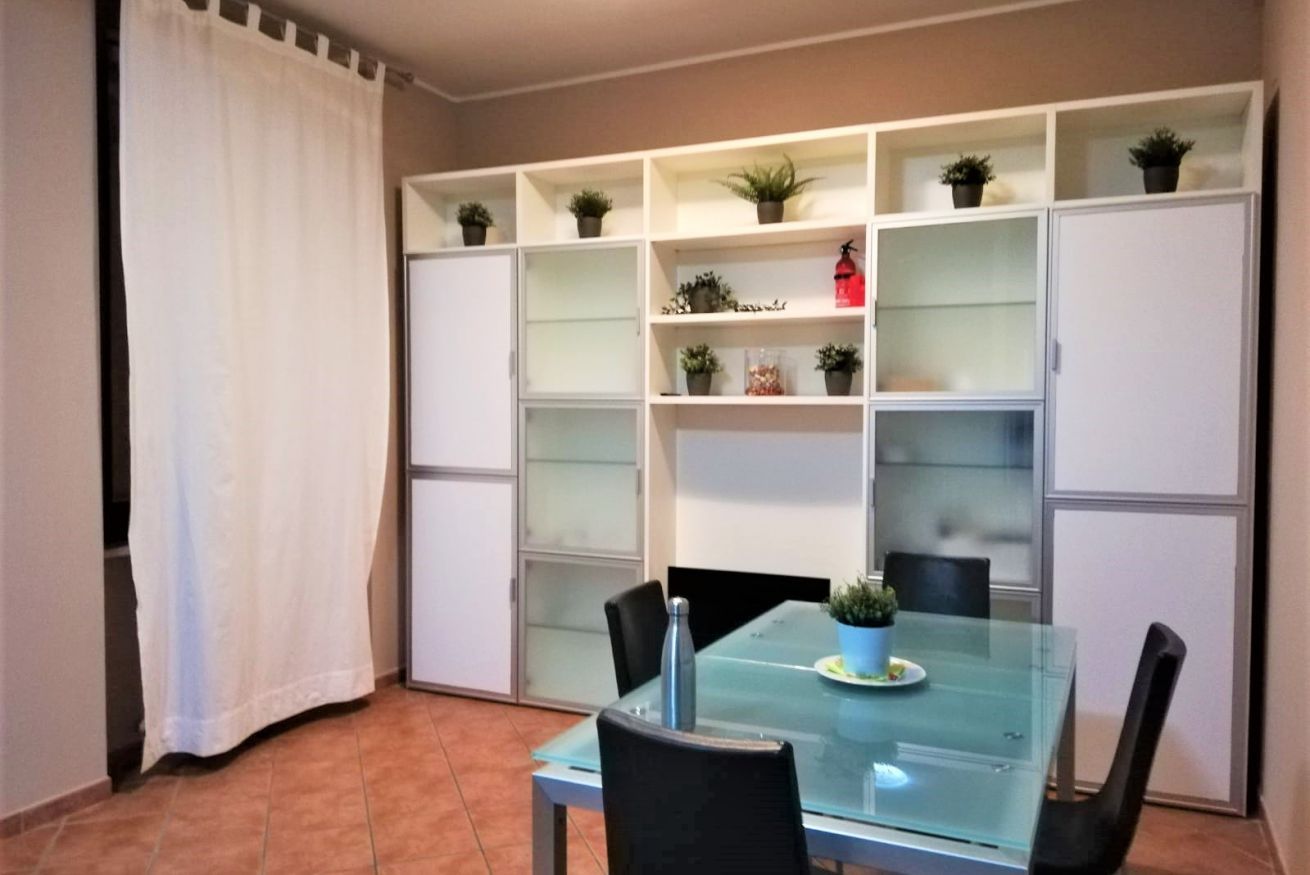 A dining area with a glass table, black chairs, and shelves with potted plants.
