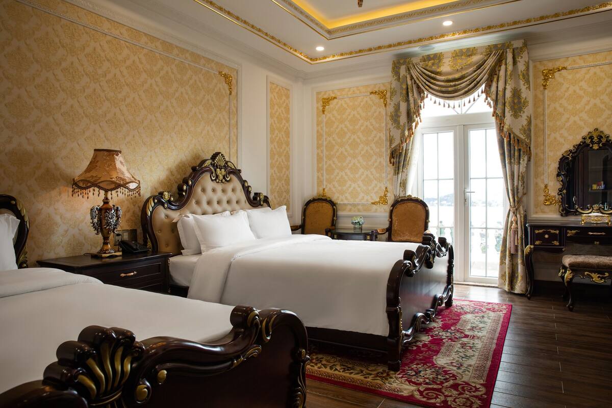 Um quarto luxuoso com duas camas grandes, cabeceiras elaboradas e um lustre clássico.