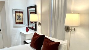 Habitación de hotel con una cama grande, dos mesitas de noche, un tocador, un espejo, un cuadro enmarcado y una ventana con cortinas.