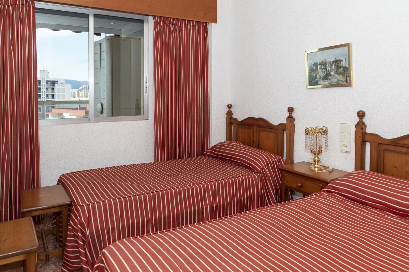 Une chambre d’hôtel avec deux lits, une fenêtre avec des rideaux, une table de chevet avec une lampe et un interrupteur fixé au mur.