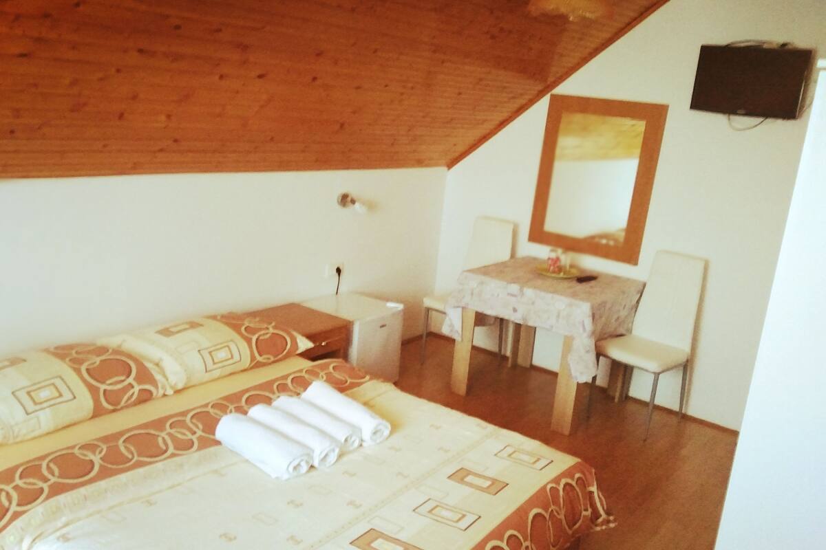 A bedroom with a wooden ceiling, a bed with patterned bedding, a desk with a chair, and a mirror on the wall.