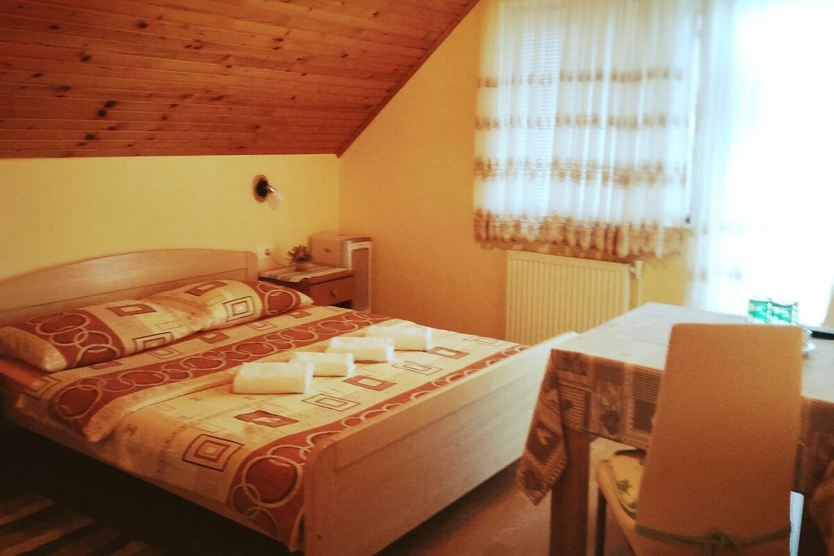 A bedroom with a wooden ceiling, a bed with patterned bedding, a bedside table, a chair, and a window with curtains.