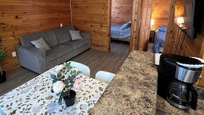 Interior acolhedor de uma cabana com decoração em madeira, um sofá cinza, uma mesa de jantar com toalha de mesa florida e uma cafeteira.