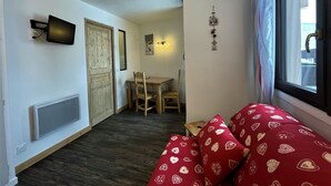 Et lille, hyggeligt rum med trægulv, en rød sofa med hjertermønster, et spisebord med stole, et fjernsyn ophængt på væggen og et vindue med gardiner.