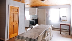 A spacious kitchen with wooden ceiling and white cabinets, a dining table with chairs, and a small window with a view.
