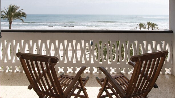 A balcony with two wooden chairs facing a beach with palm trees and the ocean.