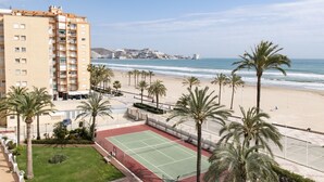 A beachfront hotel with a tennis court, palm trees, and a view of the ocean and city skyline.