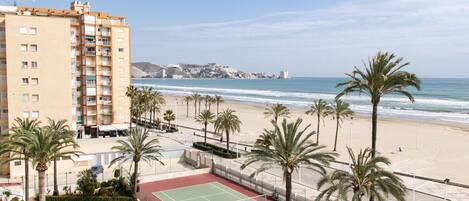 A beachfront hotel with a tennis court, palm trees, and a view of the ocean and city skyline.