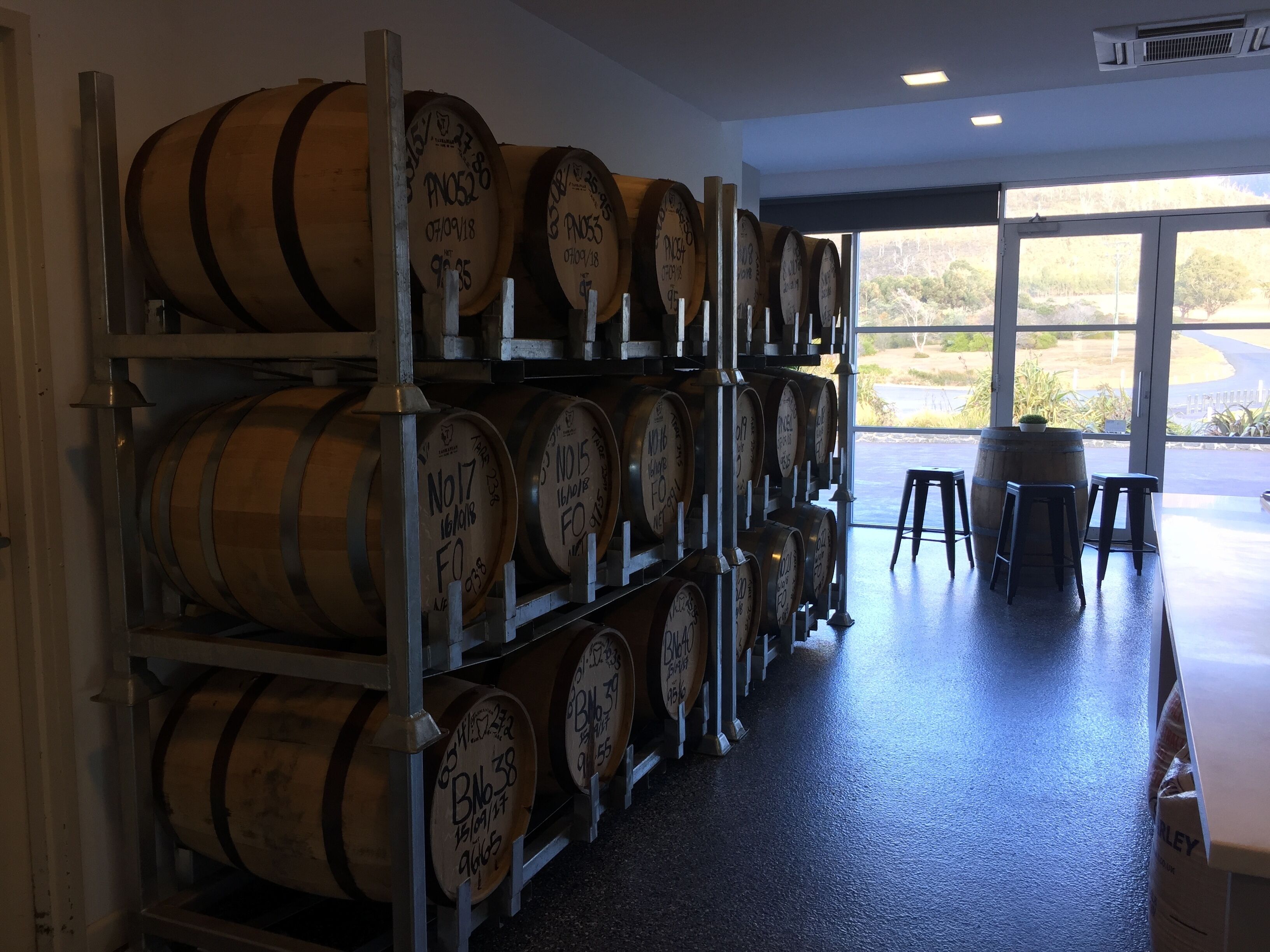 Zona de almacenamiento con barriles de madera etiquetados con números y fechas, una estantería metálica y vistas al exterior a través de un amplio ventanal.