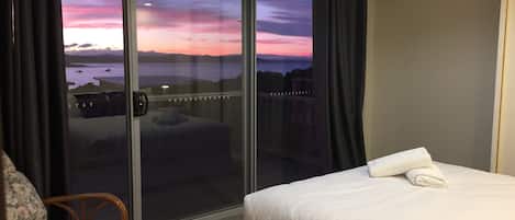 A hotel room with a bed, a chair, and a sliding glass door leading to a balcony with a view of the sea and mountains.
