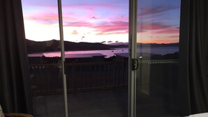 Ein Zimmer mit einem Bett, einem Sessel und einer Schiebetür zu einem Balkon mit Blick auf ein Gewässer und Hügel.