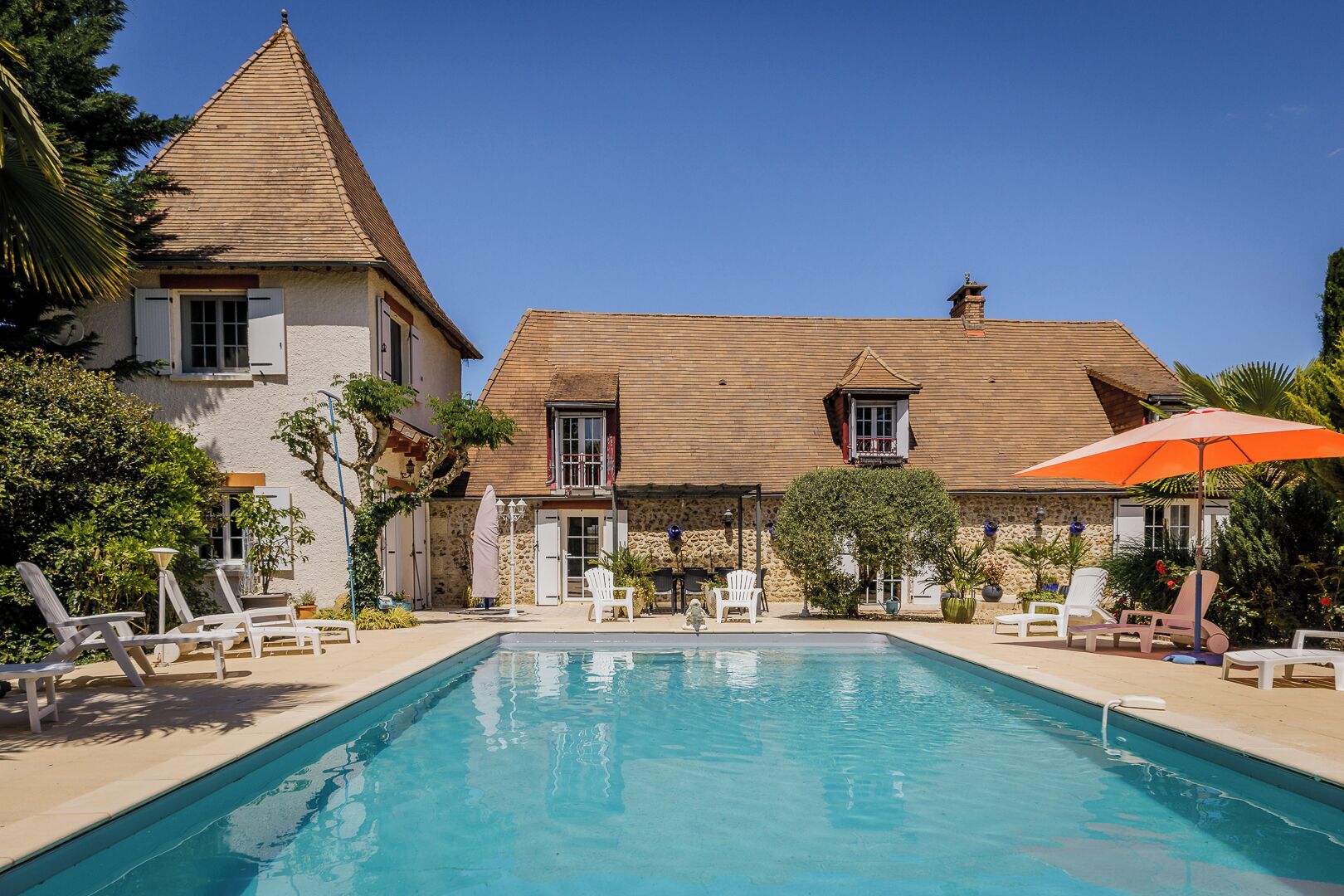Ein Poolbereich mit Liegestühlen und einem großen Sonnenschirm, umgeben von einem Steinhaus mit einem Ziegeldach und einem kleinen Turm.