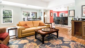 A spacious living room with a leather sofa, a coffee table, and a patterned rug. There is a dining area with a table and chairs, and a kitchen area with a red accent wall.