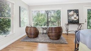 A bedroom with a large window, two wicker poufs, a bed, and a small table with a chair.