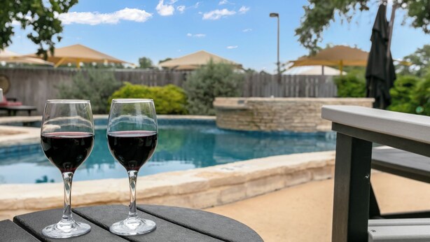 Dos copas de vino sobre una mesa junto a una piscina, con vista a un patio trasero y una valla de madera.