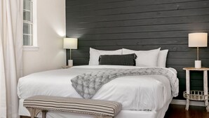 Una cama bien hecha con sábanas blancas, un plaid gris y una almohada a rayas. Hay dos mesitas de noche con lámparas, una a cada lado de la cama.