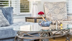 A living room with a glass coffee table, a blue sofa, a patterned armchair, a white lamp, and decorative items including a vase with red flowers and themed mugs.