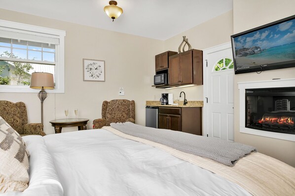 Una habitación de hotel con cama, escritorio con lámpara, kitchenette, chimenea y televisor sobre la chimenea.