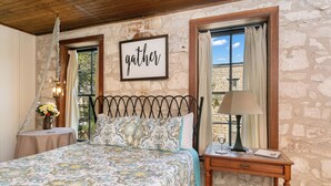 Quarto com parede de pedra, cama com roupa de cama estampada, mesa de cabeceira com abajur e janela com cortinas.