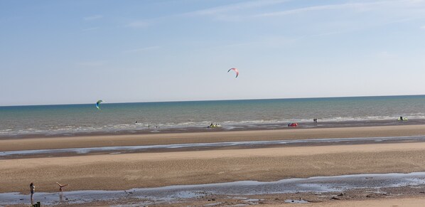 Une plage avec une vaste étendue de sable, une eau calme et quelques personnes et cerfs-volants visibles au loin.
