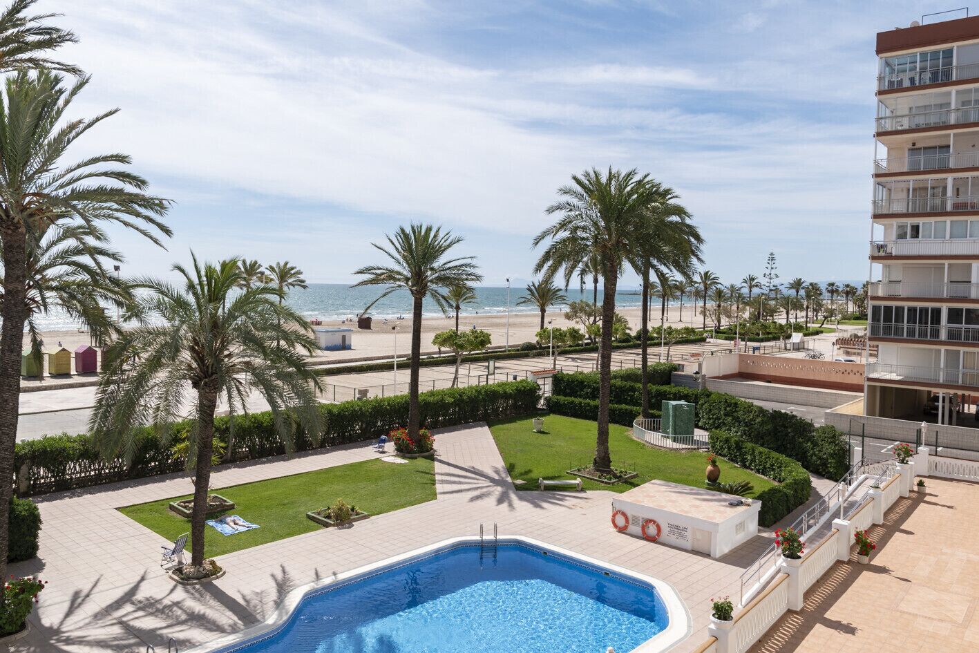 A pool area with a view of the beach and ocean, surrounded by palm trees and a multi-story building.