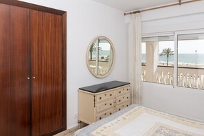 A bedroom with a wooden wardrobe, a dresser, a round mirror, and a window with a view of the beach.