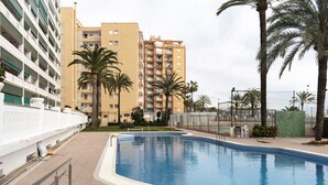 A swimming pool with a white fence, palm trees, and multi-story buildings in the background. - LIDER, 2-G (Cullera)