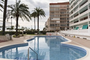 A swimming pool with a clear blue water, surrounded by a white fence, palm trees, and a multi-story building with balconies. - LIDER, 2-G (Cullera)