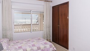 A bedroom with a bed, a wooden wardrobe, and a window with a view of a beach and a white fence. - LIDER, 2-G (Cullera)