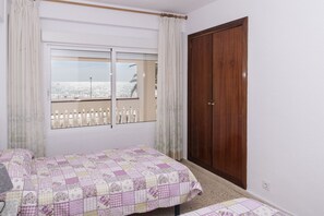 A bedroom with a bed, a wooden wardrobe, and a window with a view of a beach and a white fence.